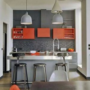 Los muebles y sillas naranjas despiertan la cocina gris.  Las paredes blancas, la isla de aluminio de estilo industrial y los accesorios de iluminación juegan con los brillantes contrastes con el gris.